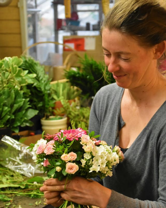 A smiling florist wrapping a bouquet of pink and cream roses in her shop, surrounded by green plants.
