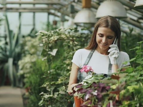 A shop owner smiling as she takes an order on her phone, surrounded by plants and flowers.